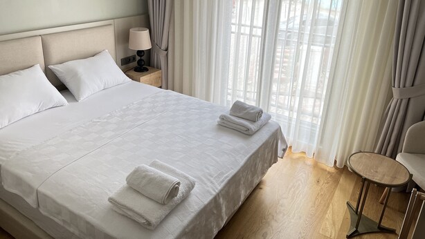 A hotel room with a large bed, two pillows, a bedside lamp, a small round table, and sheer curtains. - Sezer's Oliva Hotel (Ezine)