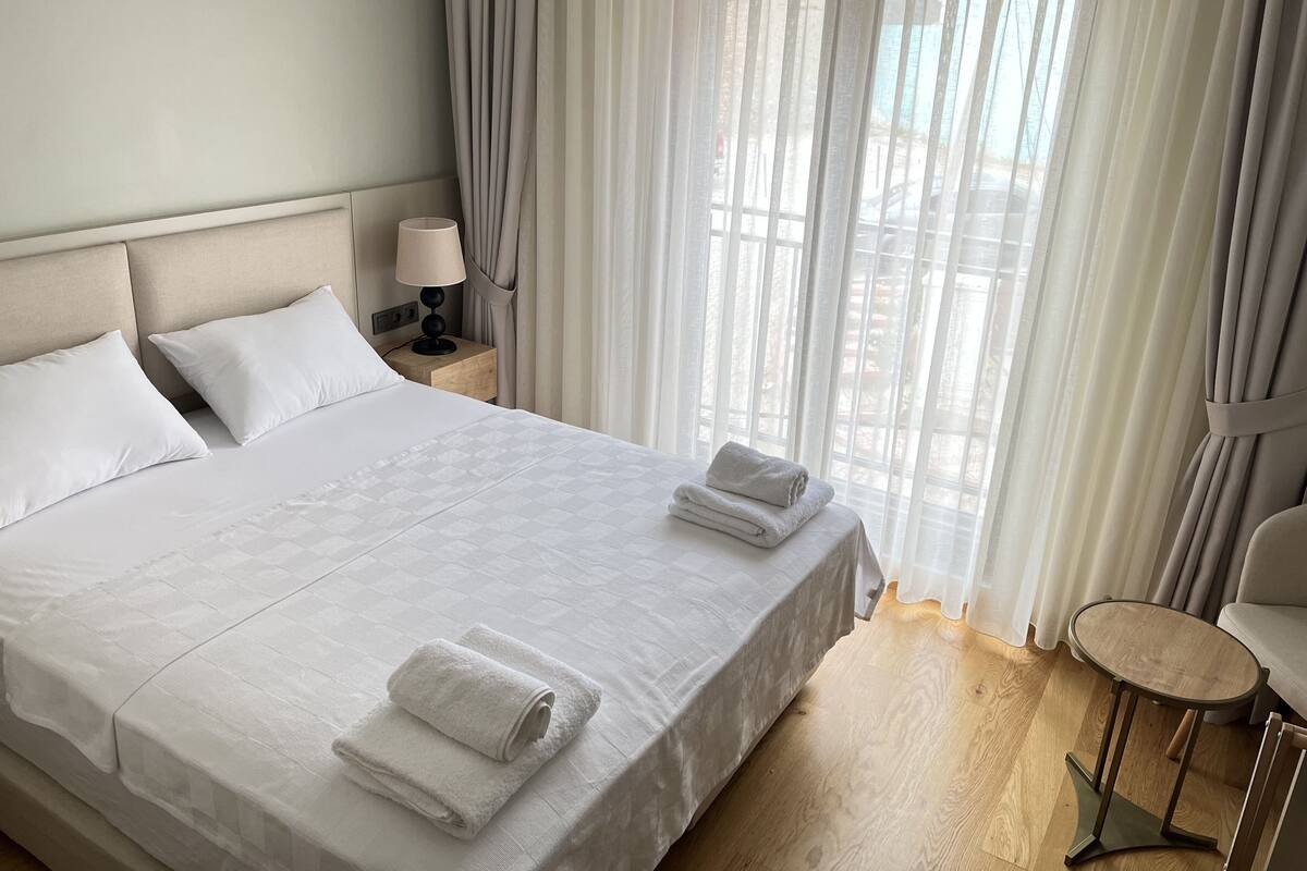 Une chambre d’hôtel avec un grand lit, deux oreillers, une lampe de chevet, une petite table ronde et des rideaux légers.
