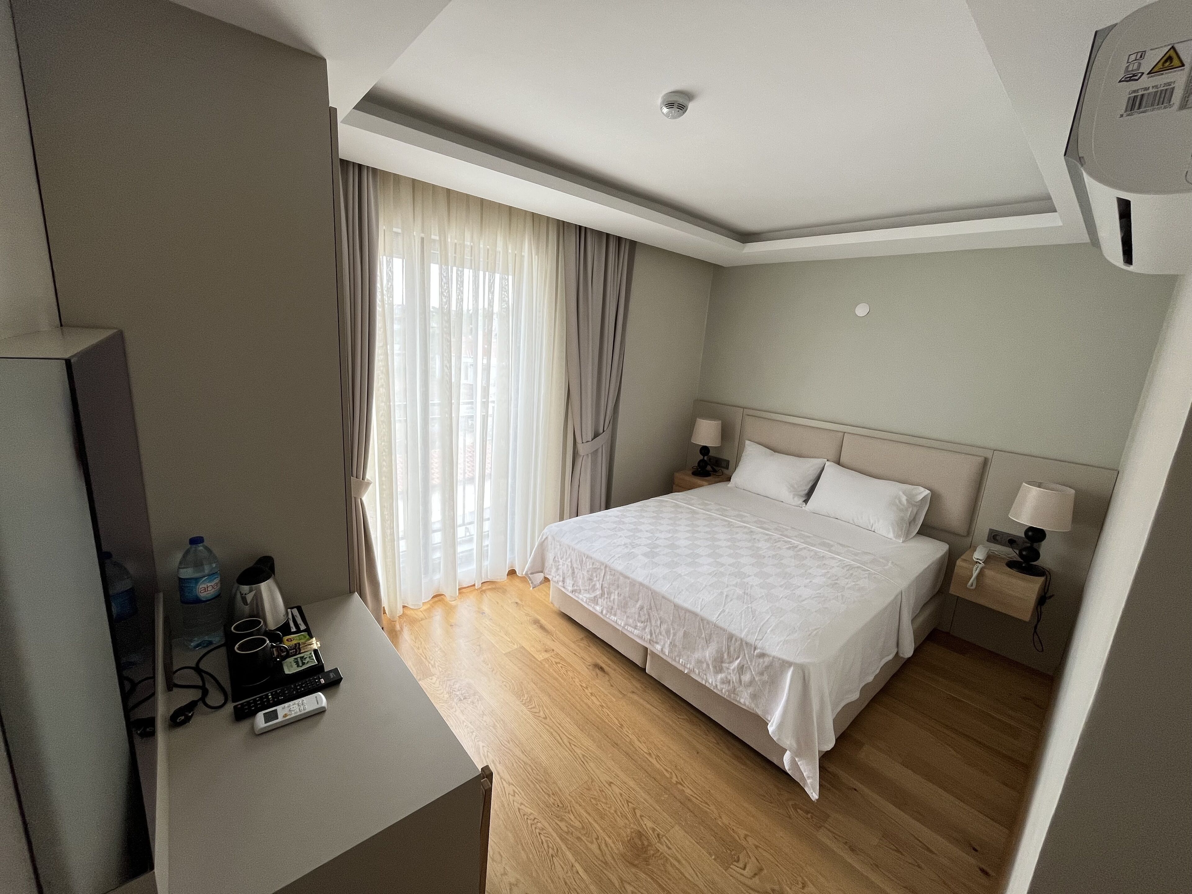 A hotel room with a bed, bedside tables, a desk with a computer and remote controls, and a window with curtains.