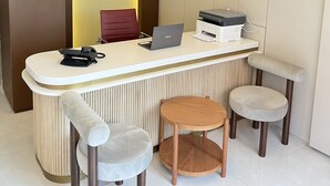 A reception desk with a laptop, a phone, and a printer. There are two cushioned chairs and a wooden stool. A security camera is mounted on the wall above the desk. - Sezer's Oliva Hotel (Ezine)