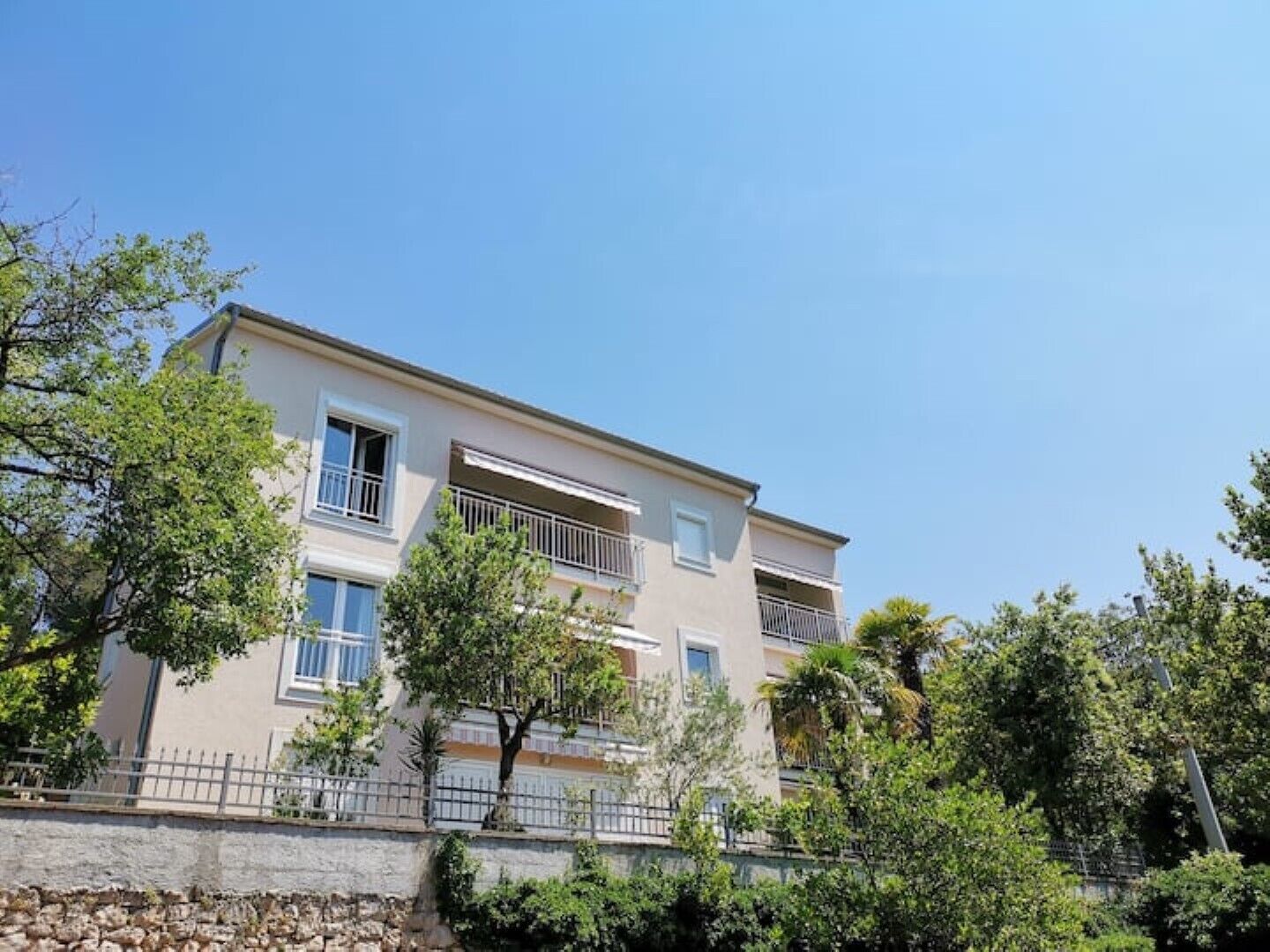 A multi-story residential building with balconies, surrounded by trees and a stone wall.
