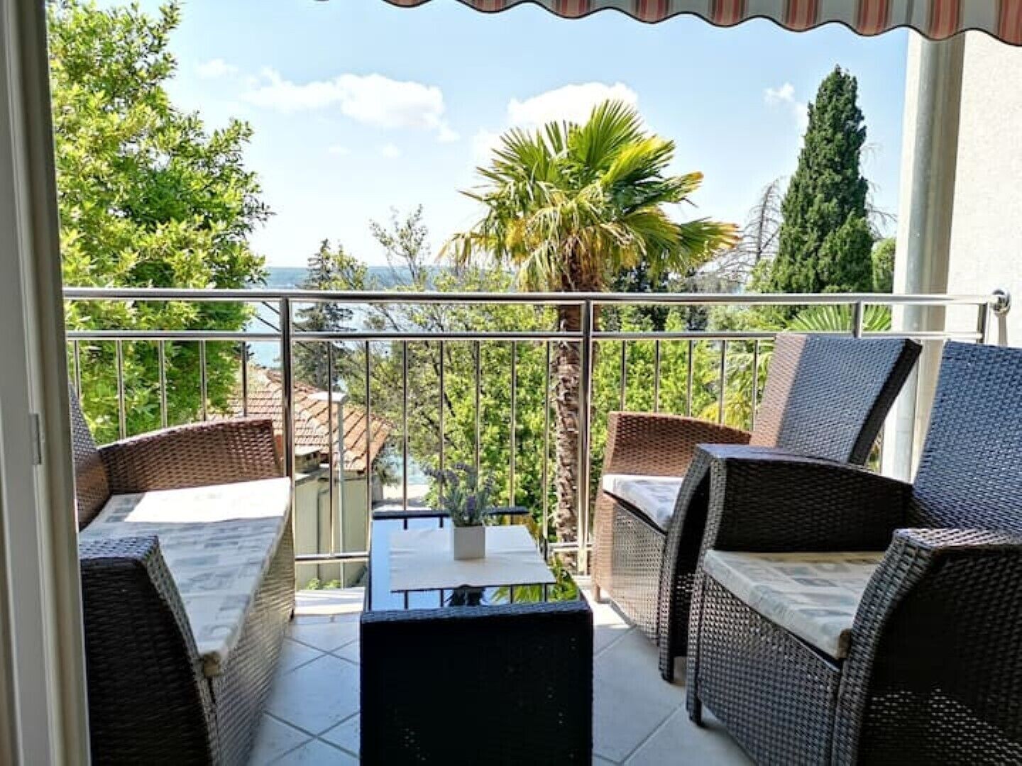 A balcony with wicker furniture, a glass table, and a view of greenery and buildings.