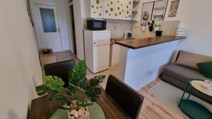 Kompakti keittiö, jossa on mikroaaltouuni, jääkaappi ja pieni keittiösaareke. Keittiössä on ruokailutila, jossa on pöytä ja tuolit, ja pöydällä on viherkasvi.