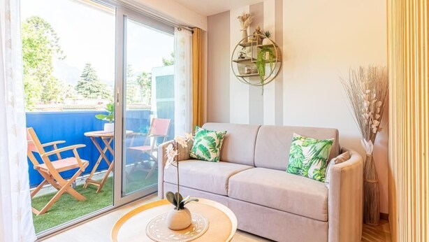 A living room with a sofa, a coffee table, and a sliding glass door leading to an outdoor area with a blue wall and wooden chairs. - Le 182 Promenade du Soleil, Tout Juste Rénové, Azur Riviera (Menton)