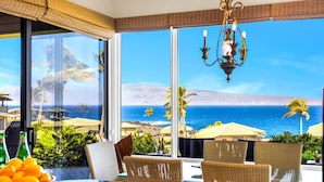 A dining area with a view of the ocean, wicker chairs, a table with a bowl of fruit, and a chandelier.