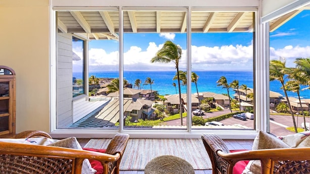 A room with a large glass door leading to a balcony with a view of the ocean and palm trees.