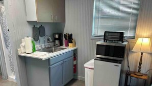 Cocina compacta con lavabo, estufa y microondas. Hay una ventana con persianas, una mesita con lámpara y un cubo de basura.