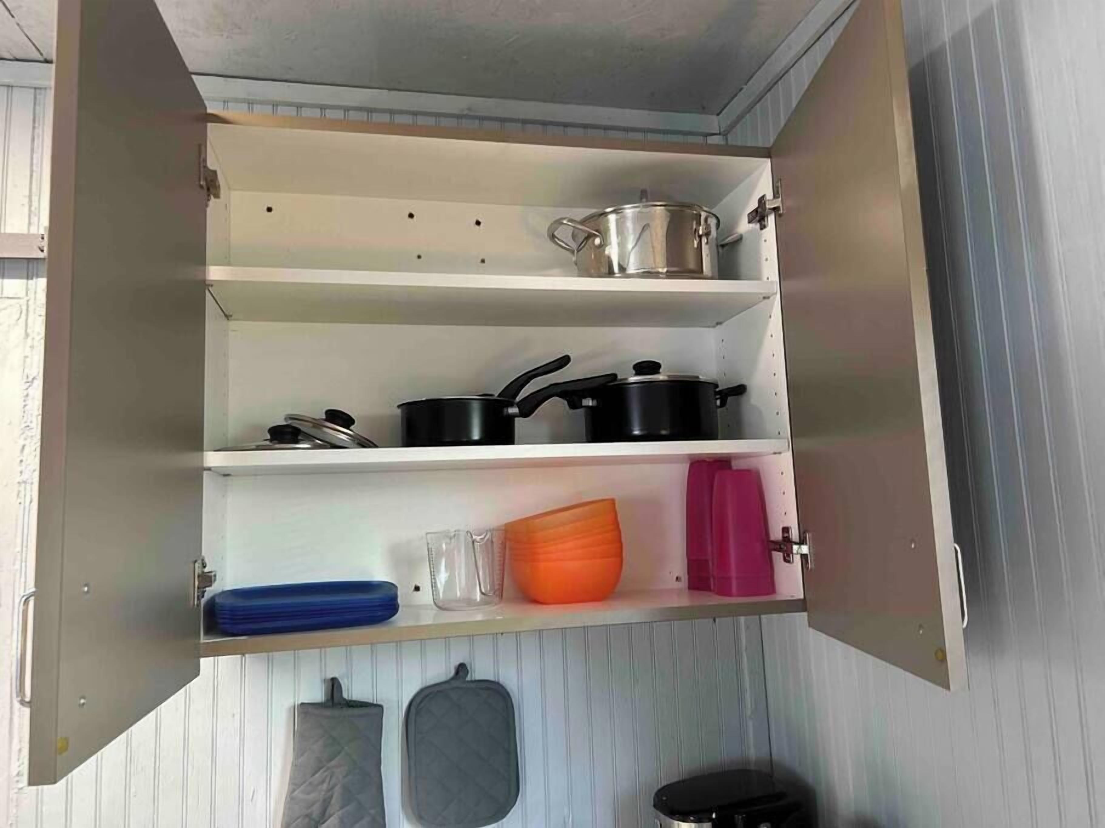 A kitchen with open cabinets, containing pots, pans, a glass, a cutting board, and a kettle.