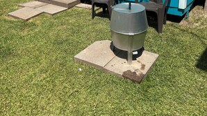 Una casa azul con puerta y ventanas blancas, dos sillas de plástico y una parrilla metálica sobre una plataforma de concreto.