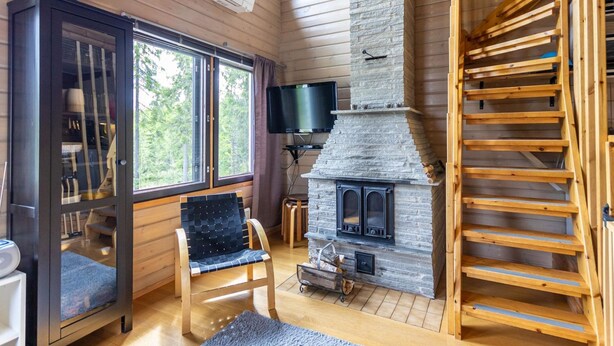 A cozy cabin interior with a stone fireplace, wooden floors, a comfortable chair, and a large window offering a view of the forest. - Kuusipirtti by Interhome (Puolanka)