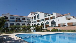 A modern hotel with a large swimming pool, multiple balconies, and a clear blue sky.