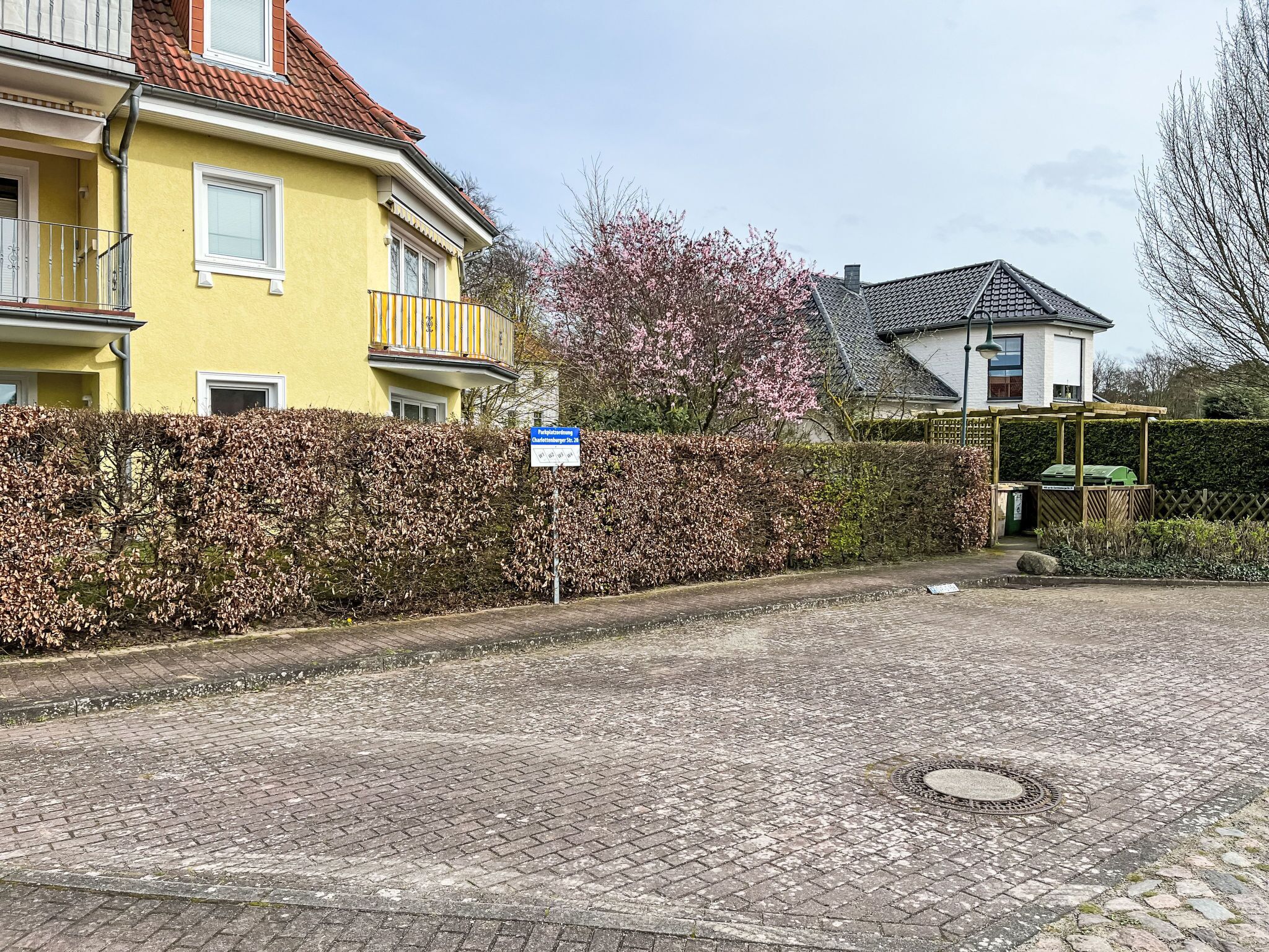 A cobblestone driveway leading to a yellow house with a red roof, surrounded by a hedge and a tree with pink blossoms.