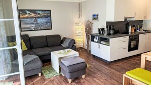 Una sala de estar moderna con un sofá gris, una mesita de centro blanca pequeña y un otoman gris. Se observa un área de cocina con gabinetes blancos y una estufa negra.