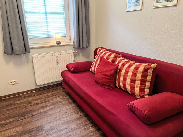 Un canapé rouge avec des coussins à rayures, un radiateur, une fenêtre avec des rideaux et un mur orné de deux tableaux encadrés.