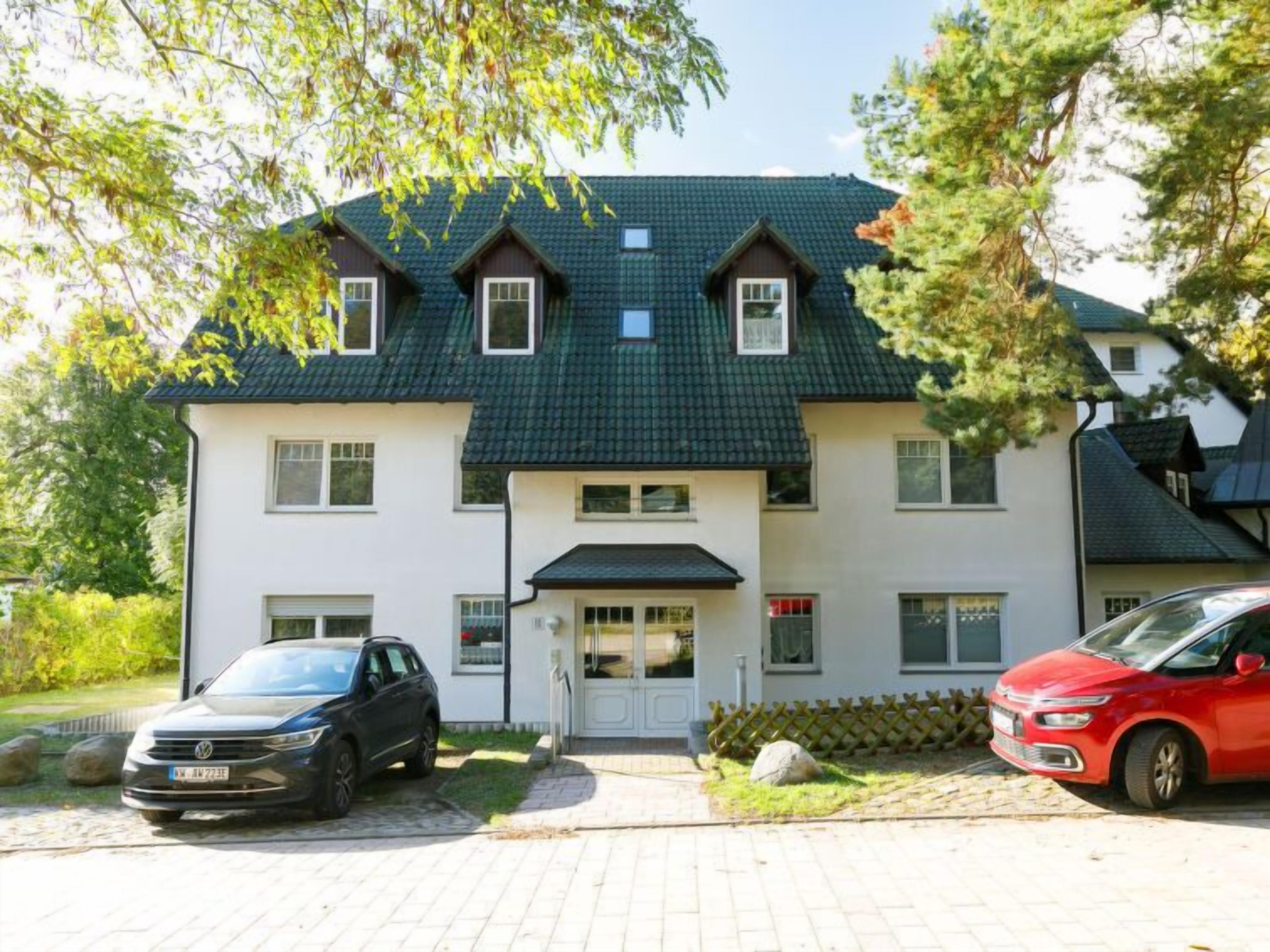 A two-story house with a dark green roof, a white facade, and a front entrance with a small porch. There are two cars parked in the driveway.