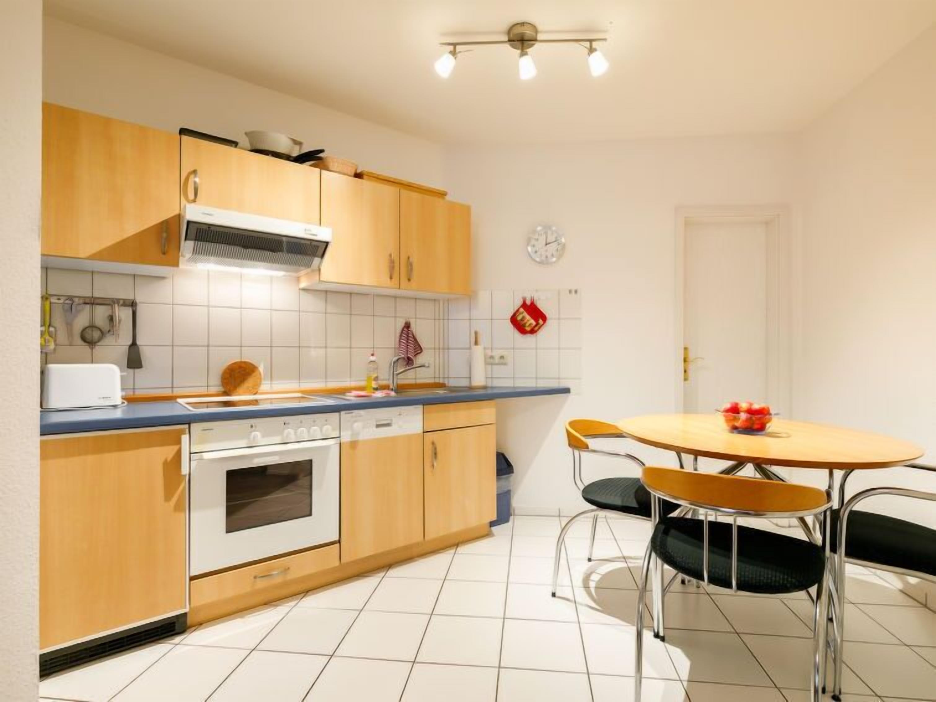 A compact kitchen with wooden cabinets, a built-in oven, a dishwasher, and a round dining table with two chairs.