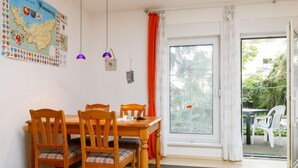 Un coin repas avec une table et des chaises en bois, une carte accrochée au mur et, à travers une porte vitrée, une vue sur un espace extérieur aménagé pour s’asseoir.
