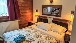 Une chambre avec un mur en bois, un lit recouvert d’une literie à motifs floraux, une table de chevet avec une lampe et un tableau accroché au mur.