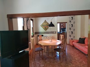 Uma sala de jantar com mesa e cadeiras de madeira, uma televisão em primeiro plano e uma cozinha ao fundo.