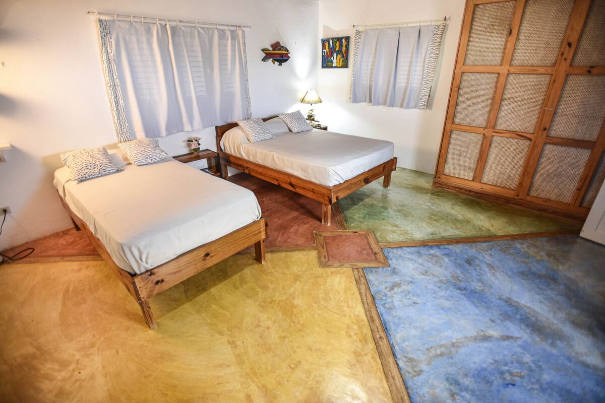 A room with two wooden beds, a small table with a lamp, and a large wooden door.
