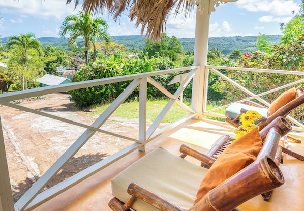 Una terraza de madera con barandilla blanca, una silla de lounge con cojín y vistas a un paisaje exuberante con árboles y una cadena montañosa distante.
