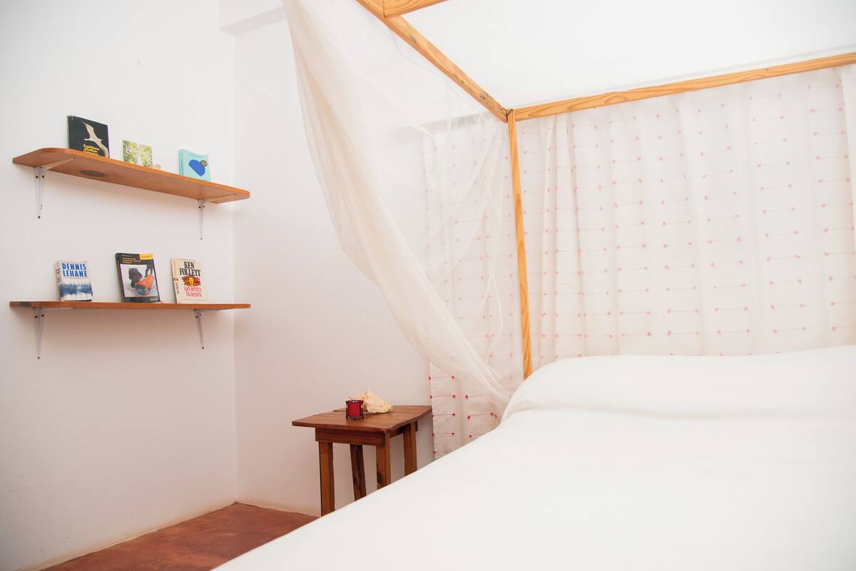 A room with a bed, a wooden nightstand, a small table, and a shelf with books and a candle.