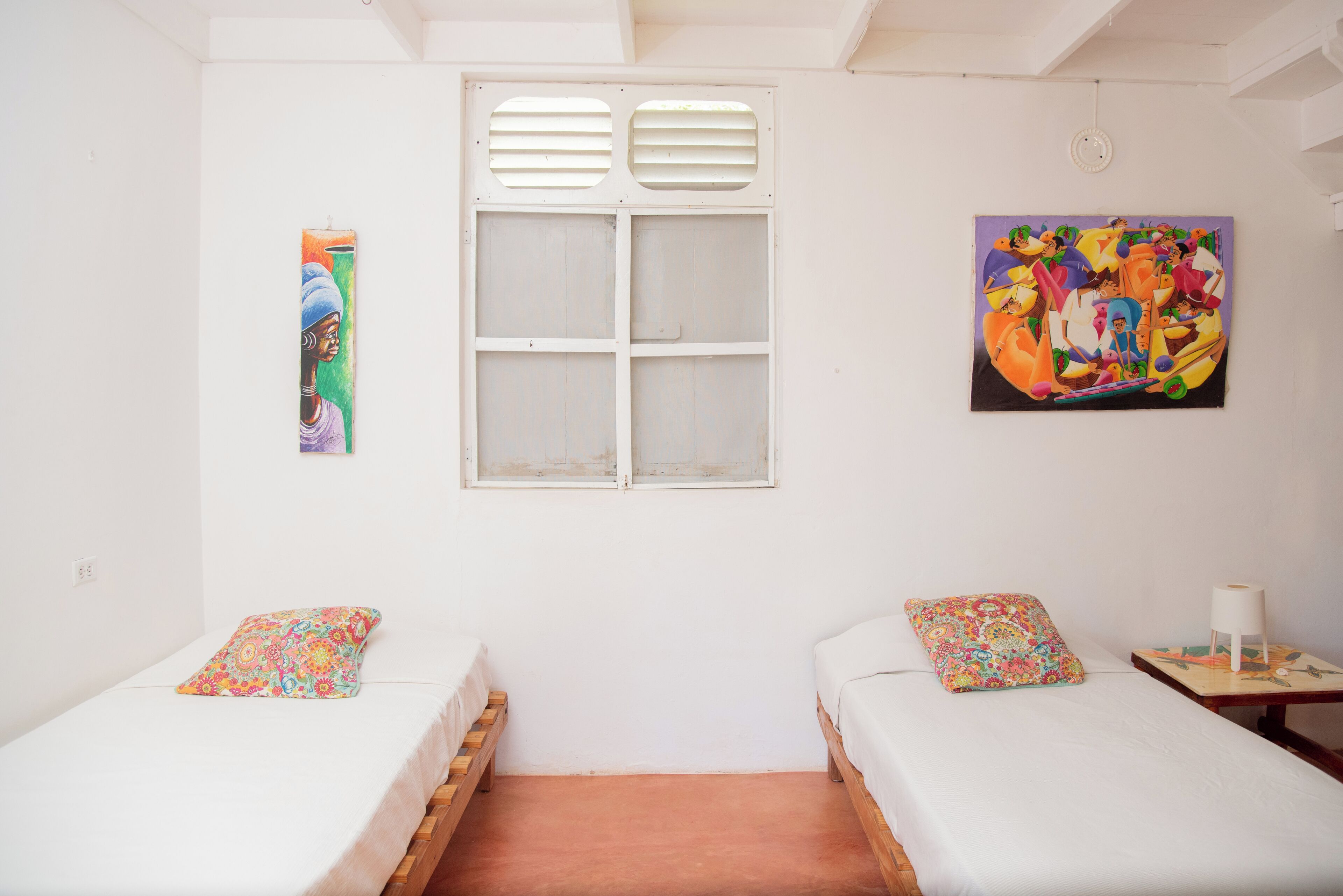 Une chambre avec deux lits simples, une fenêtre avec des volets, une peinture murale et une petite table avec une lampe.