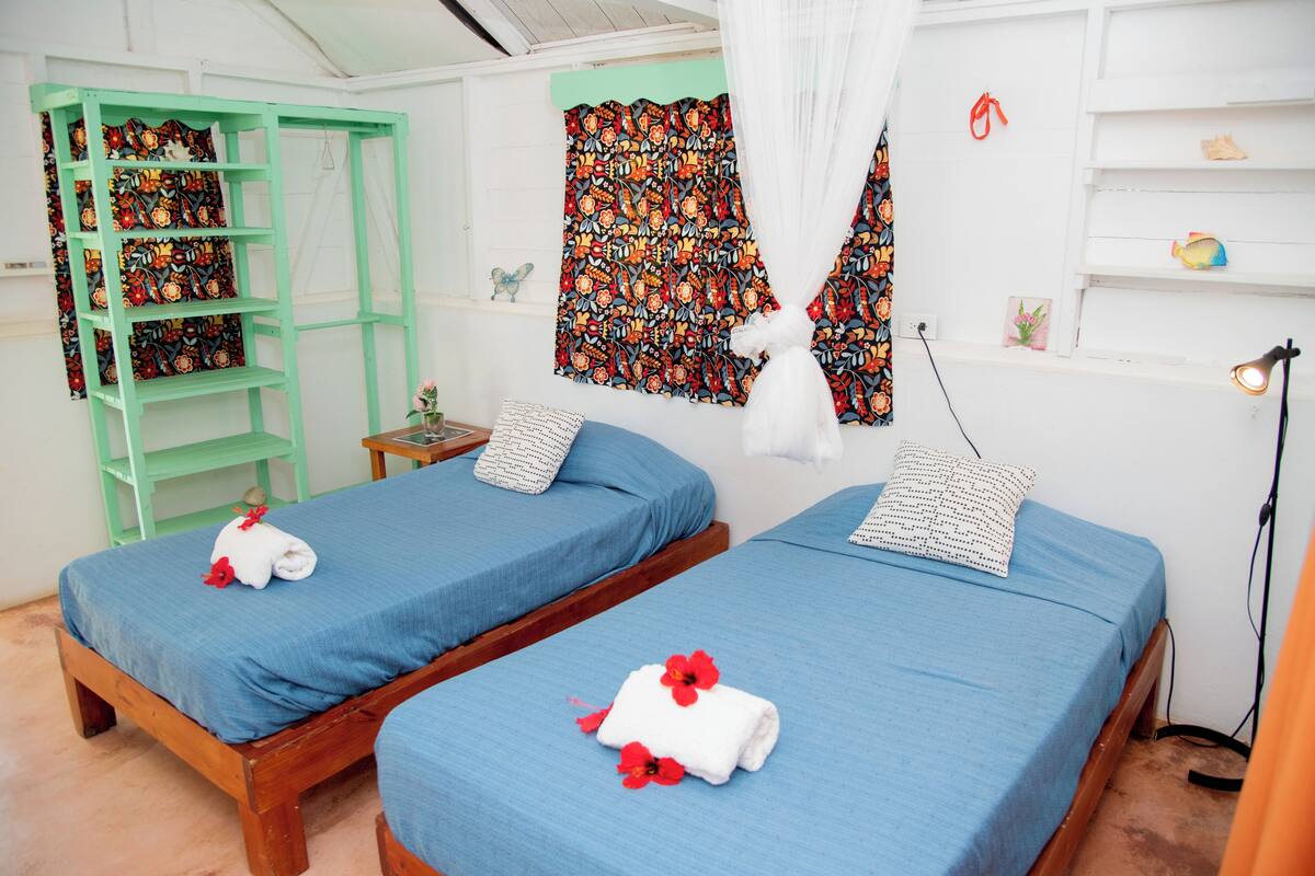 A room with two single beds, a green shelf unit, a patterned curtain, and a small table with a lamp.