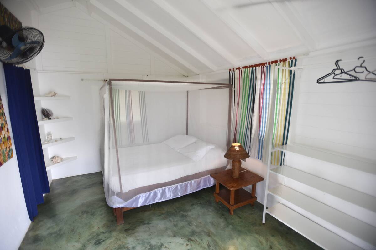 A single-bed room with a canopy bed, a bedside table with a lamp, a shelf with decorative items, and a wall-mounted coat rack.