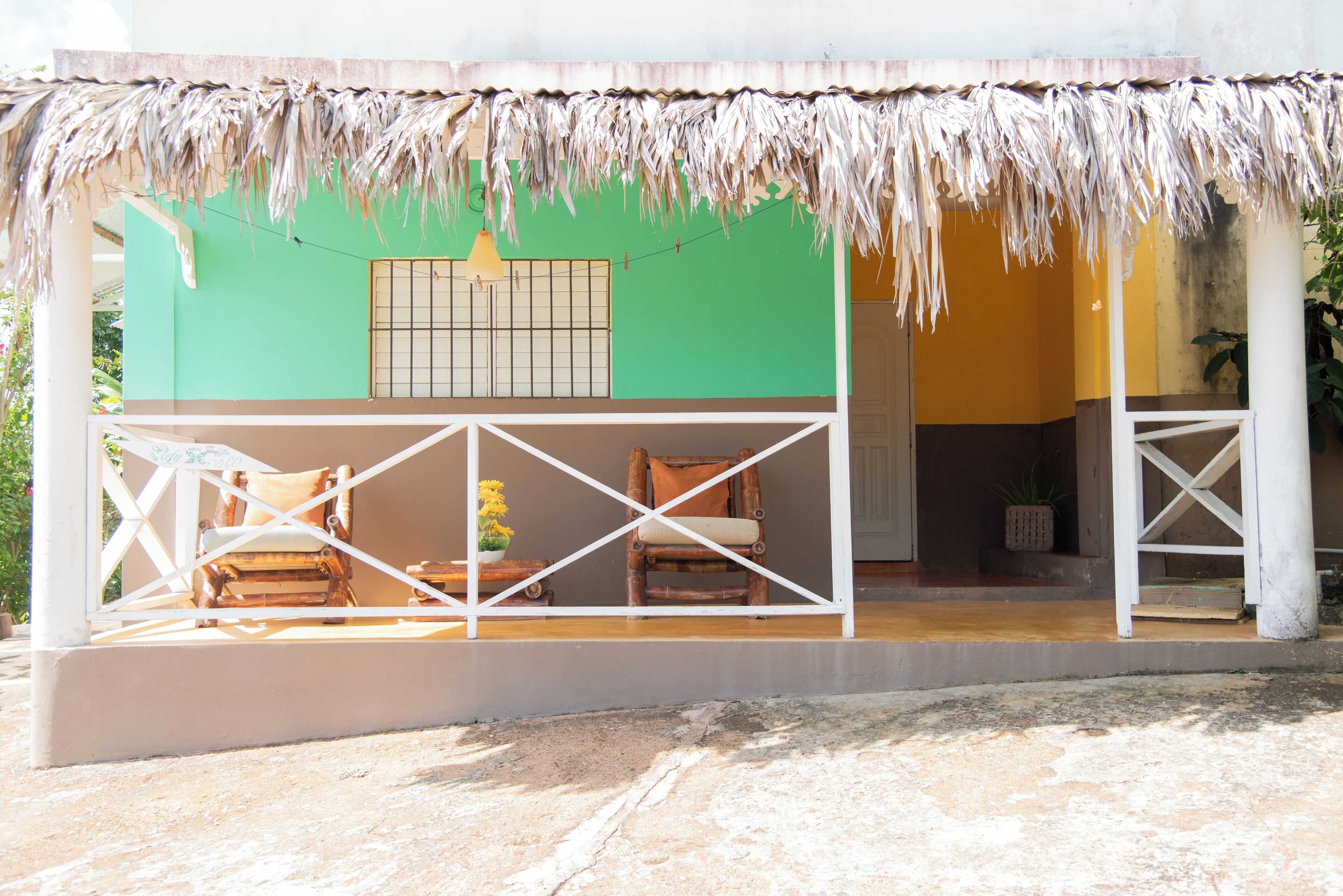 A thatched-roof structure with a green and yellow wall, two wooden chairs, and a potted plant.