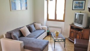 Una sala de estar con un sofá gris, una mesita con un florero, un televisor sobre un mueble de madera y una ventana con cortinas.