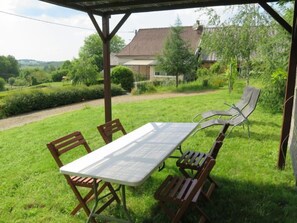 A covered patio with a table and chairs, surrounded by lush greenery and a view of a house in the background. - Rillettes by Interhome (Pompadour)