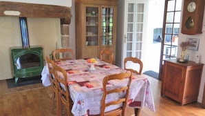 Un comedor con una mesa de madera, sillas, mantel acolchado, estufa de leña verde, un mueble de madera y un reloj de pared.