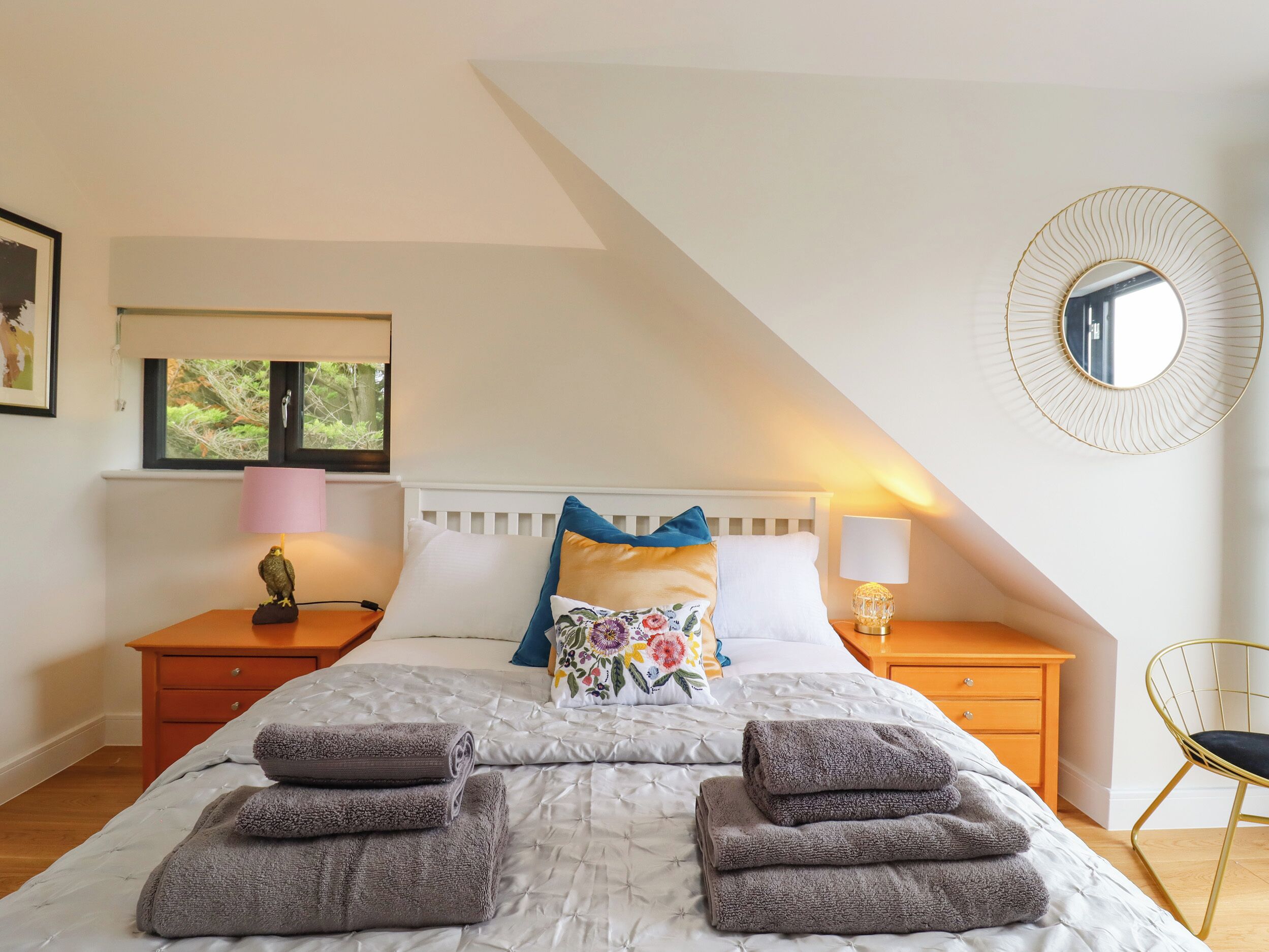 A bedroom with a bed, pillows, a nightstand, a lamp, a mirror, and a window.
