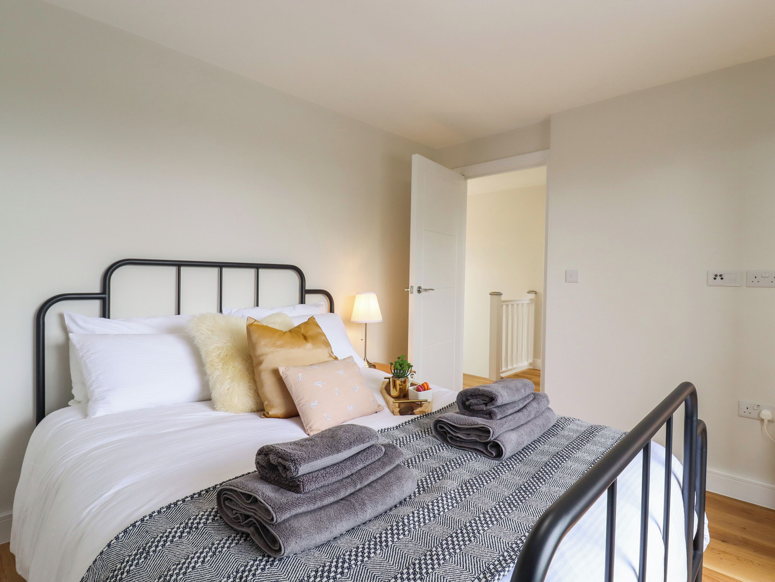Un lit bien fait, avec des oreillers blancs et beiges, une tête de lit en métal noir et une couvre-lit à motifs. On peut voir des serviettes pliées sur le lit, ainsi qu’une petite table de chevet sur laquelle se trouvent une lampe et une plante.