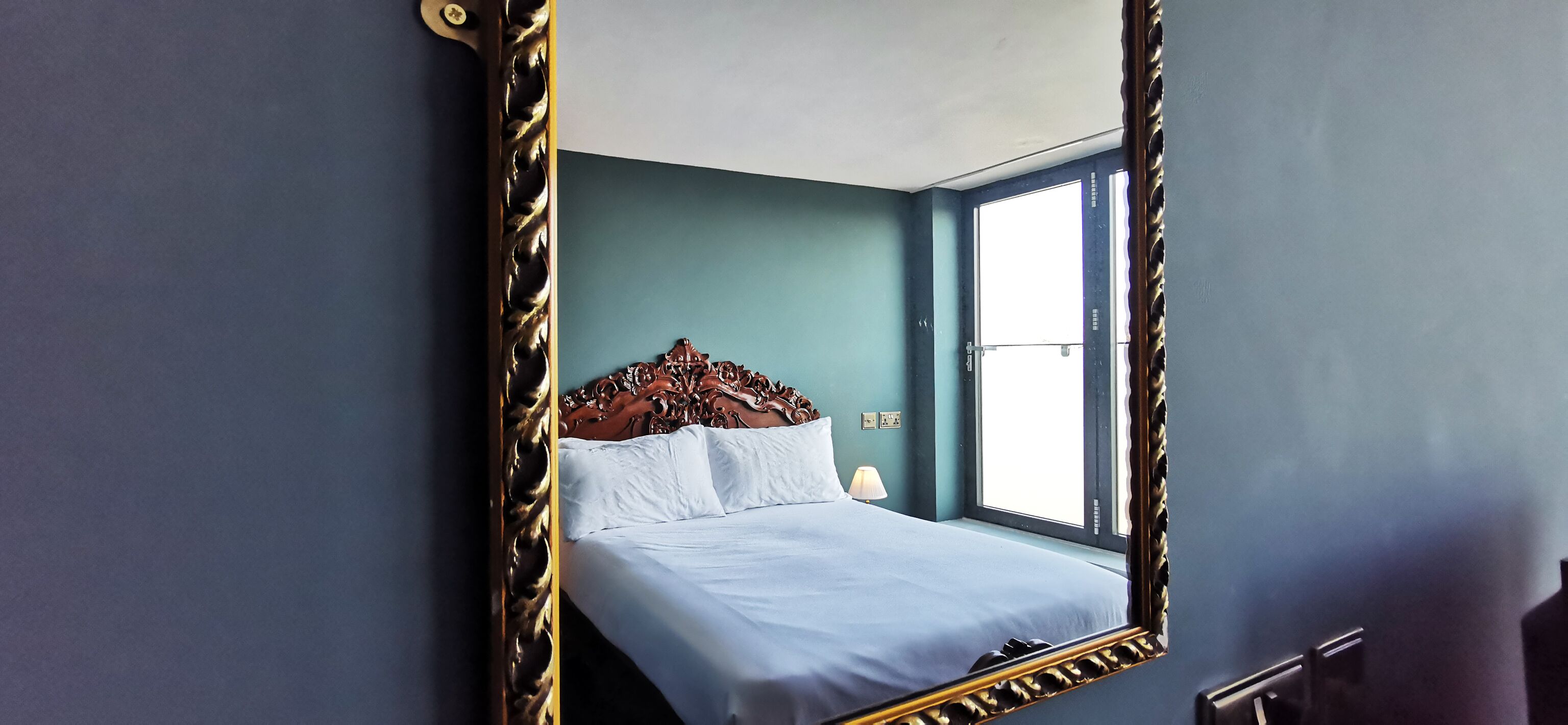 A bedroom with a large ornate mirror, a bed with white linens, a wooden headboard, and a window with daylight.