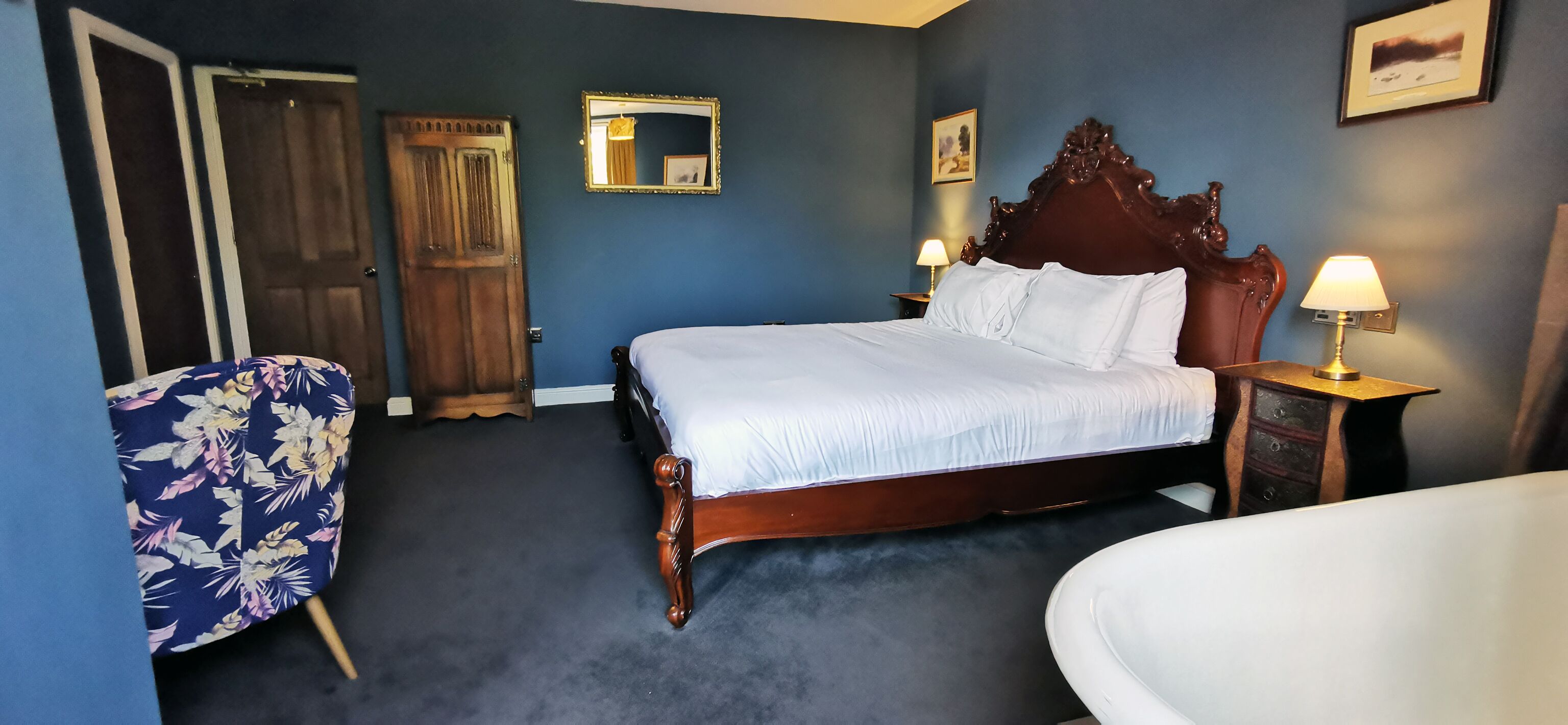 Ein Hotelzimmer mit einem großen Bett, zwei Nachttischen mit Lampen, einem Holz-Kopfteil, einem blumigen Sessel und einer Tür.
