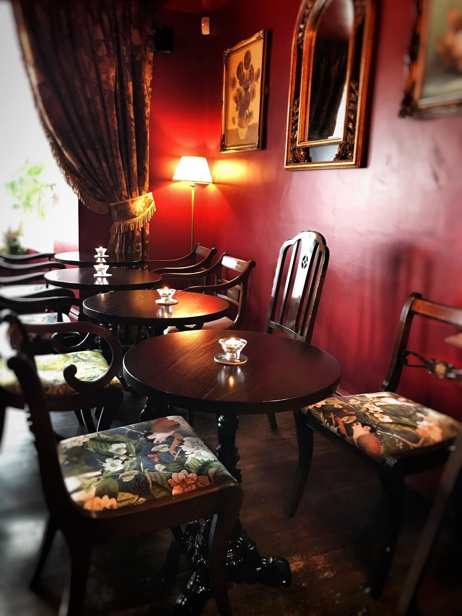 A dimly lit restaurant with round tables and chairs, a lamp, and framed artwork on the walls.