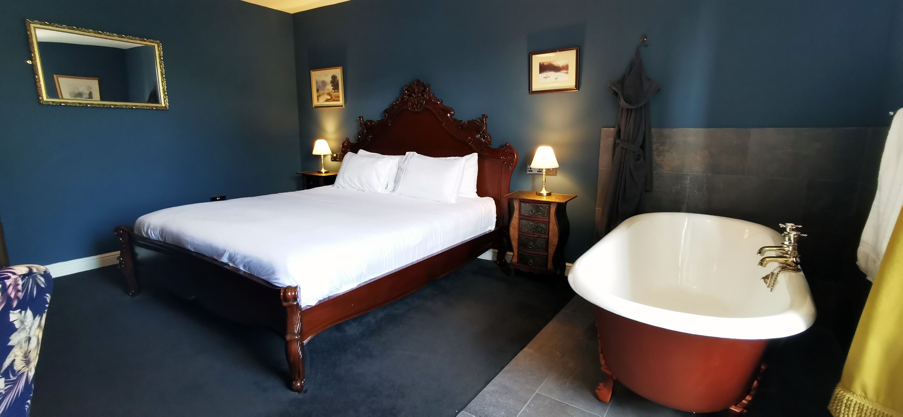 A bedroom with a large bed, a wooden headboard, two bedside tables with lamps, and a large freestanding bathtub.