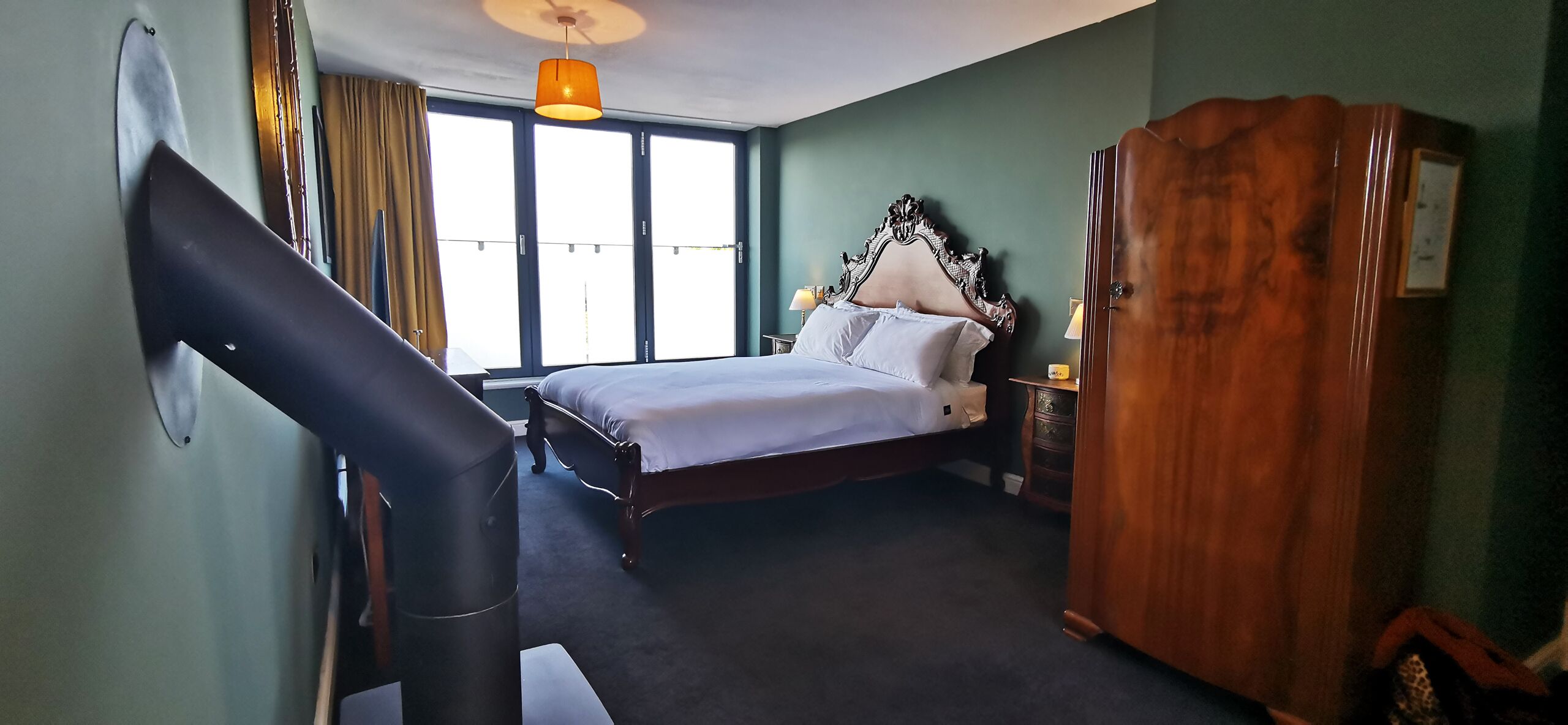 Hotellihuone, jossa on sänky, puinen vaatekaappi, lamppuvarustettu työpöytä ja suuri ikkuna, jossa on verhot.