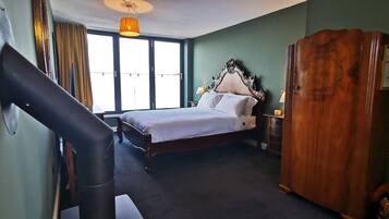 A hotel room with a bed, a wooden wardrobe, a desk with a lamp, and a large window with curtains.