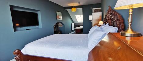 A bedroom with a large bed, a wooden headboard, a bedside table with a lamp, a TV mounted on the wall, and a skylight.