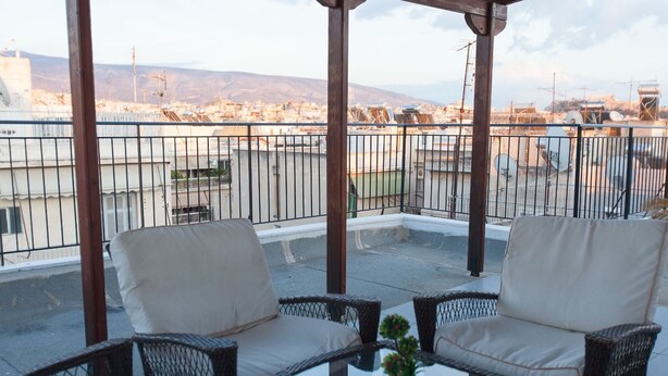 A covered outdoor seating area with wicker furniture and a glass table, overlooking a cityscape with buildings and mountains. - Acropolis view! (Athina)