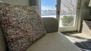 A room with a large window, a sofa with a map-patterned pillow, and a view of a snowy landscape. - The Alex Furnished Rentals (Oshkosh)