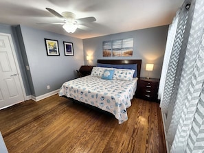 Quarto com cama, ventoinha no teto, mesa de cabeceira, abajur, quadros nas paredes e janela com vista.