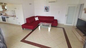 Ein Wohnzimmer mit einem roten Sofa, einem Couchtisch und einem Fernseher. Im Hintergrund ist eine Küche mit weißen Schränken und einer Mikrowelle zu sehen.