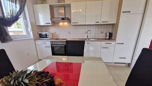 Eine moderne Küche mit weißen Schränken, einer Mikrowelle, einem Herd und einem Spülbecken. Ein Esstisch mit rotem Tischläufer und einer Obstschale mit Bananen, Trauben und einem Apfel.
