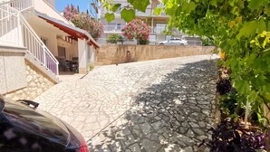 Un carrer pavimentat amb un cotxe aparcat a l'esquerra, un edifici amb una escala i un balcó amb flors roses.