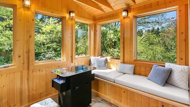 L’intérieur d’un chalet en bois est aménagé avec un banc intégré et une petite table. De grandes fenêtres offrent une vue sur la verdure. À l’extérieur, on trouve un banc en pierre.