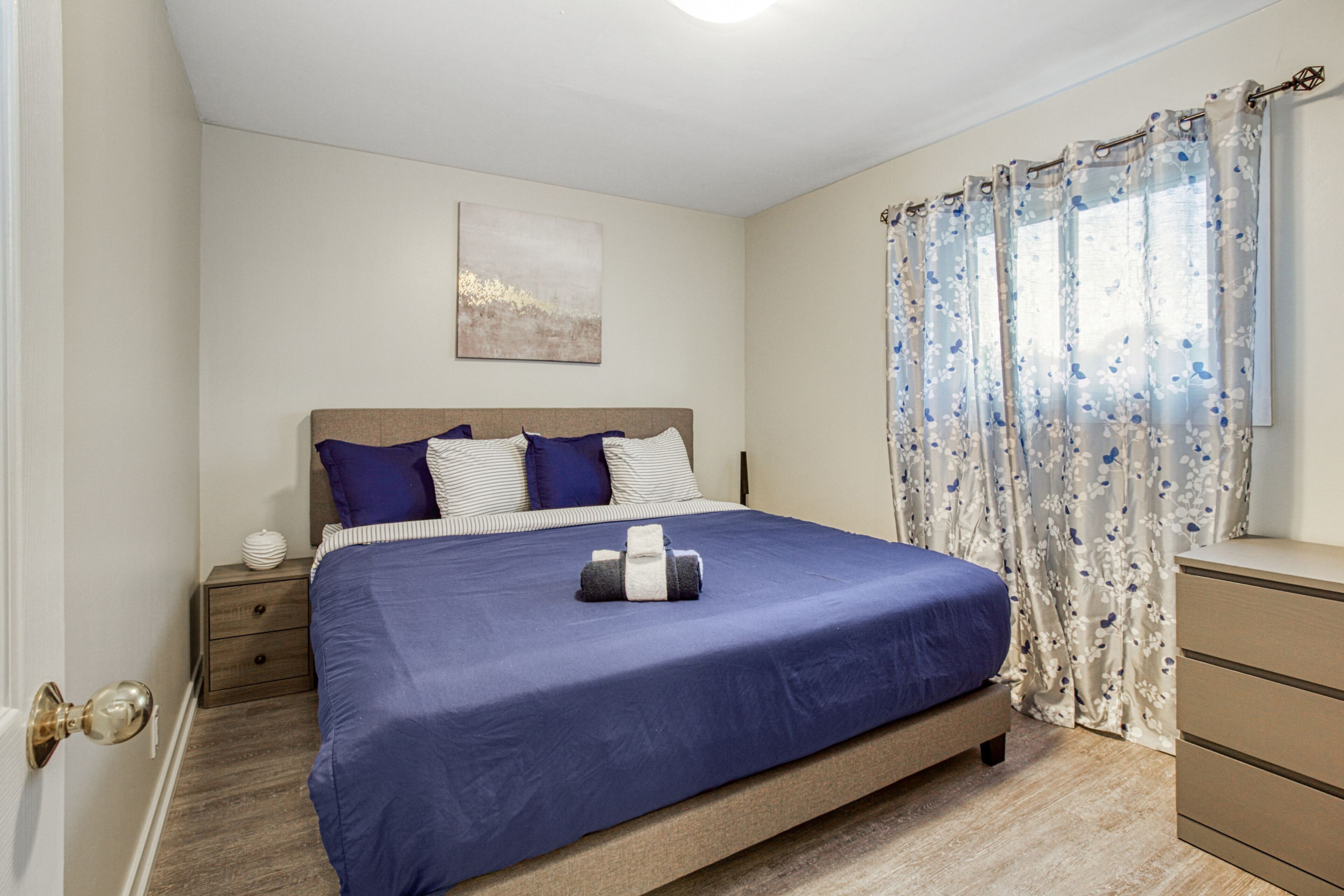 Een slaapkamer met een bed, nachtkastje, dressoir, raam met gordijnen en een aan de muur hangend kunstwerk.